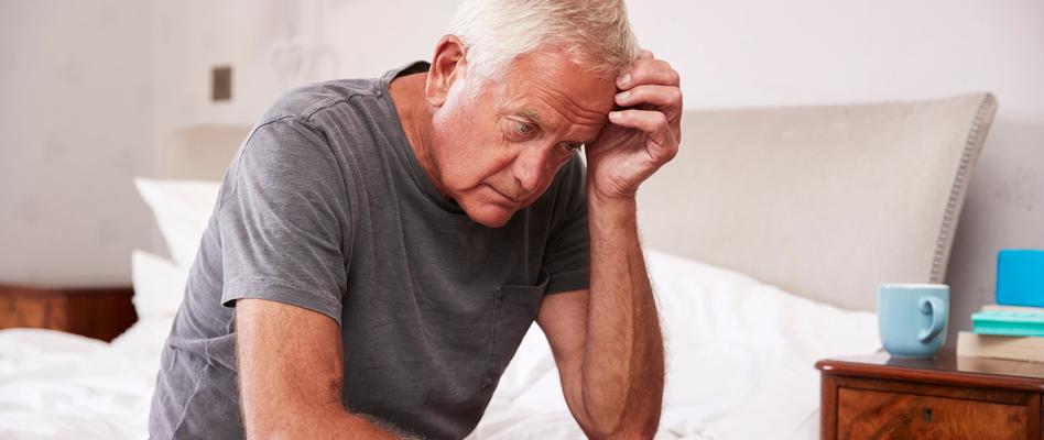 A man is sitting on the edge of his bed wearing a grey sleep shirt. He has a hand on his head and appears to be confused or dazed. He is staring at the ground. 