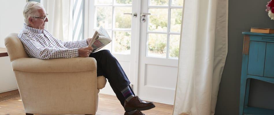 An elderly gentleman is sitting in a tan arm chair with his legs crossed. He is reading the newspaper. He is sitting by a set of french doors that lead outside. 