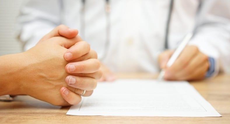 A pair of hands is on the left side of the image, crossed on a wooden table. A doctor is reviewing paperwork to the left of the hands. The doctor is holding a pen, nudging to writing on the paperwork in front of them.