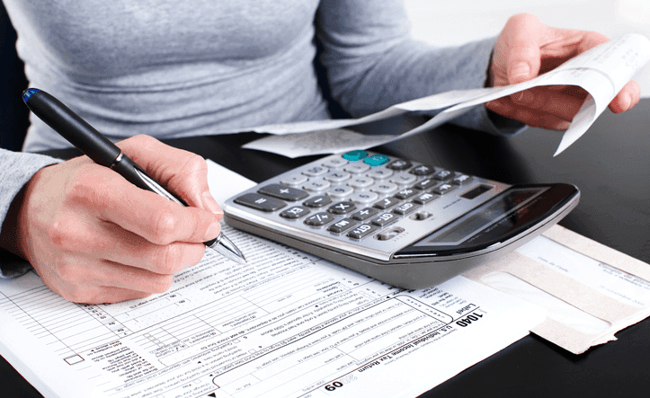 A woman is sitting at a desk with tax paperwork. She has a calculator in front of her and is holding a pen. 